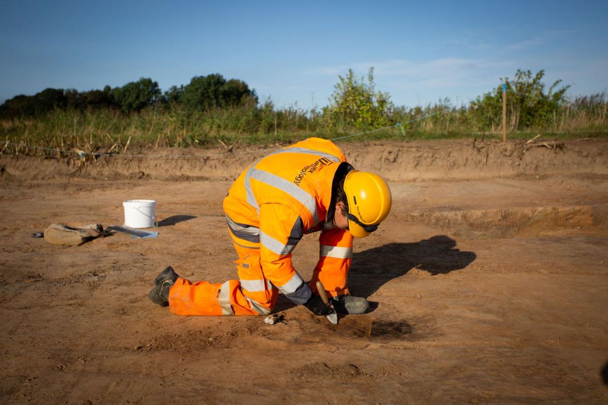 An archaeologists crouches with one knee extended, using a trowel to carefully remove earth from an artefact.
