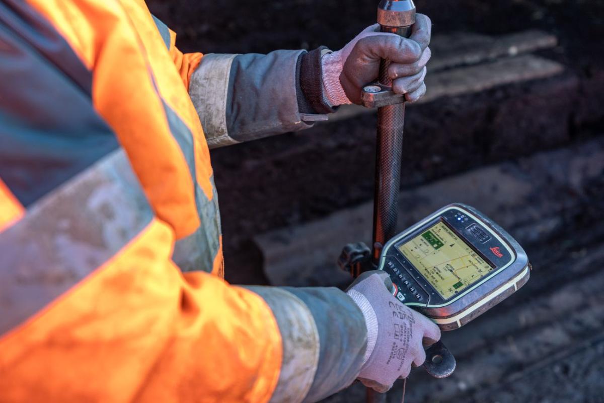 A field archaeologist is stood in the trench, holding a measuring stick.