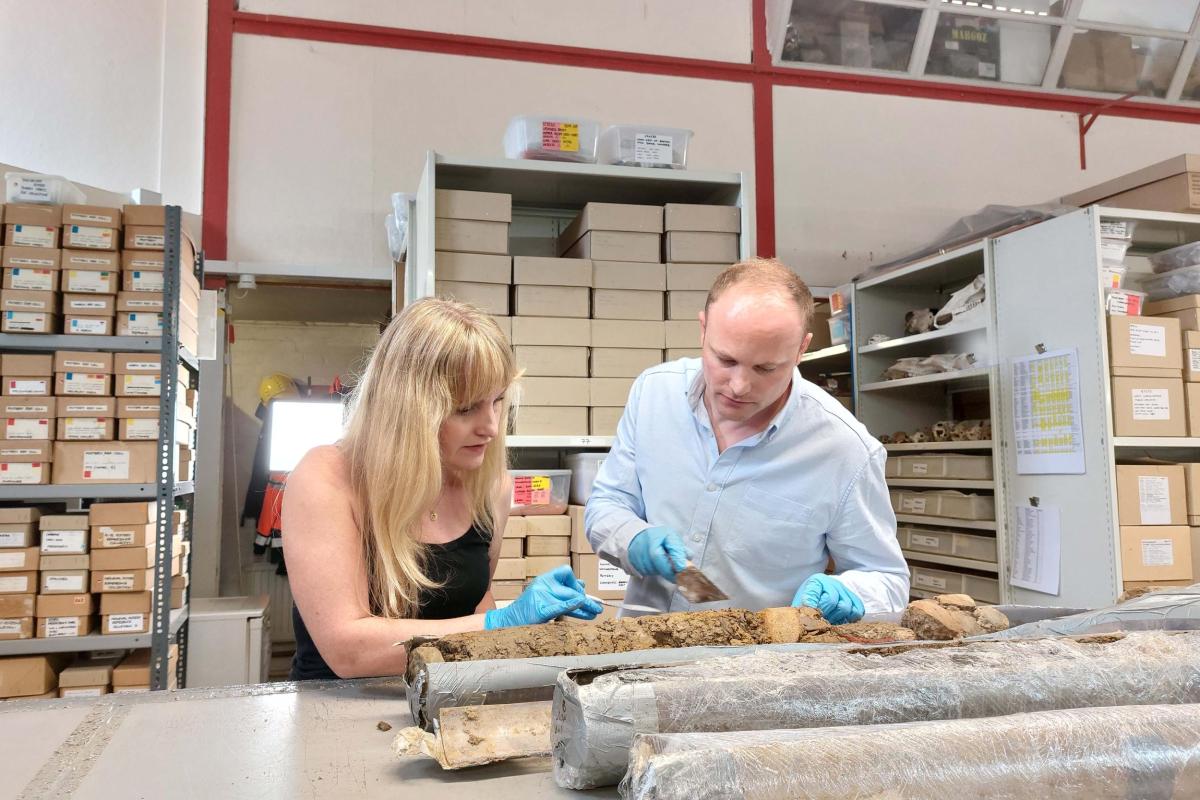 Image of Dr Cat Jarman and Dr Daniel Young. Both are wearing blue gloves. Dr Daniel Young is holding a trowel to show parts of the Sheffield Castle samples which are placed on a bench in front. 