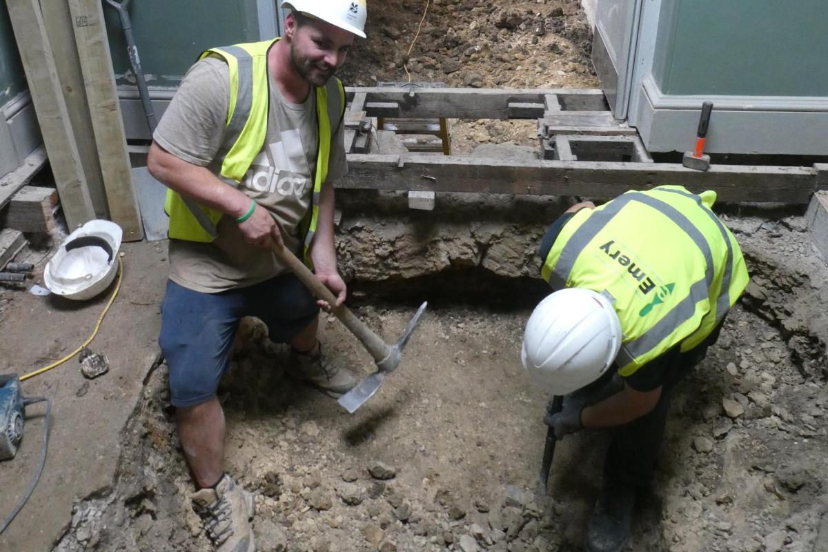 Two archaeologists in hard-hats and high vis are shown digging up the floor beneath Bath Assembly Rooms with pick-axes.