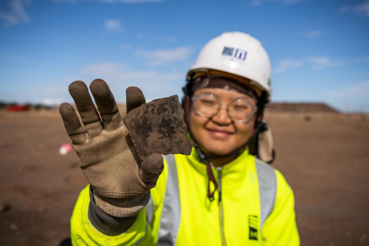 A field archaeologist smiles at the camera and holds up a black piece of pottery.