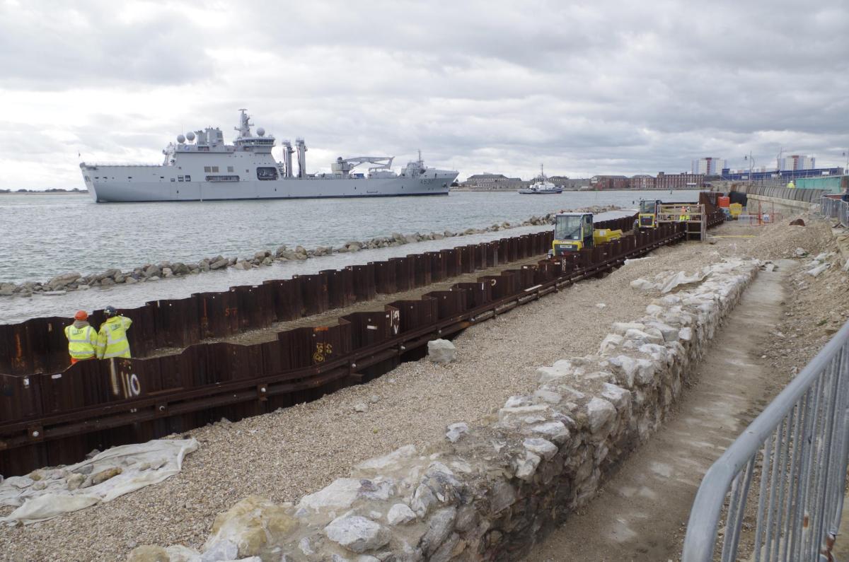 Southsea Coastal Scheme defences made out of stone gabions and metal shoring extends along the coast, the sea glimmers and large modern ship floats on the horizon