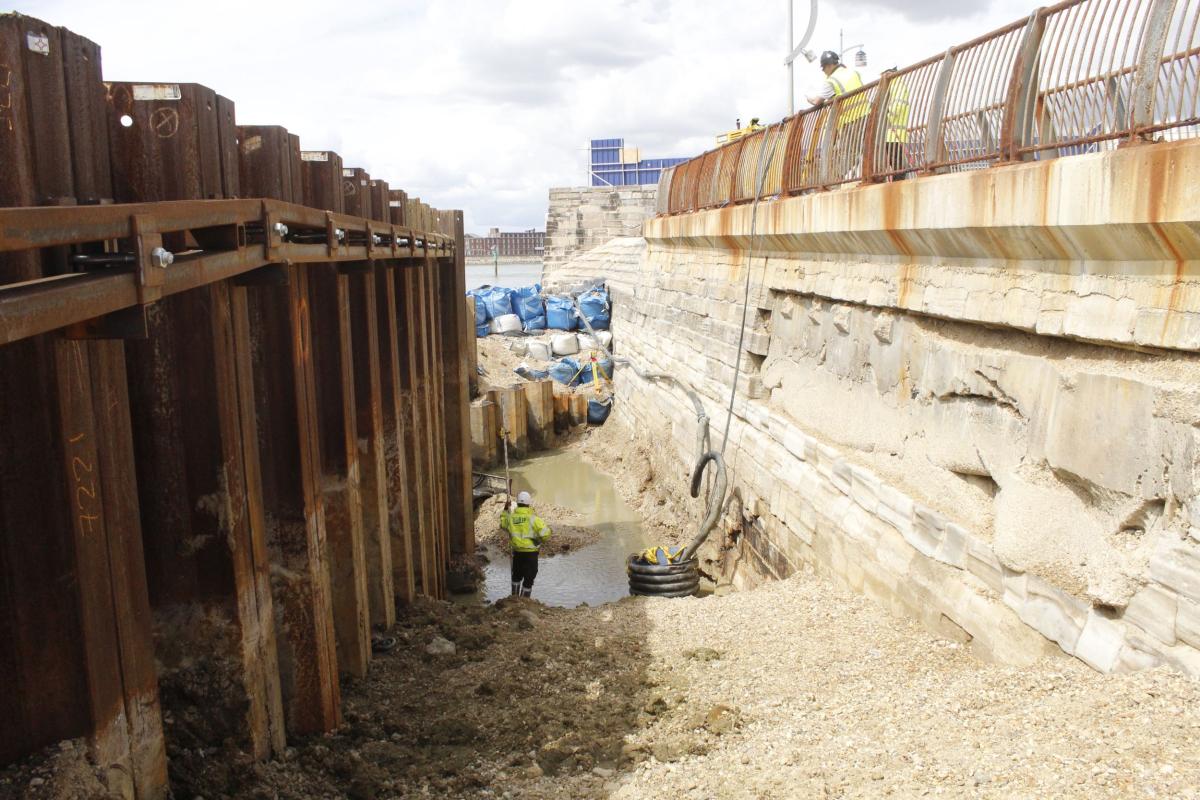 Old and new sea defences run parallel at the water's edge, a construction worker stares at the tide