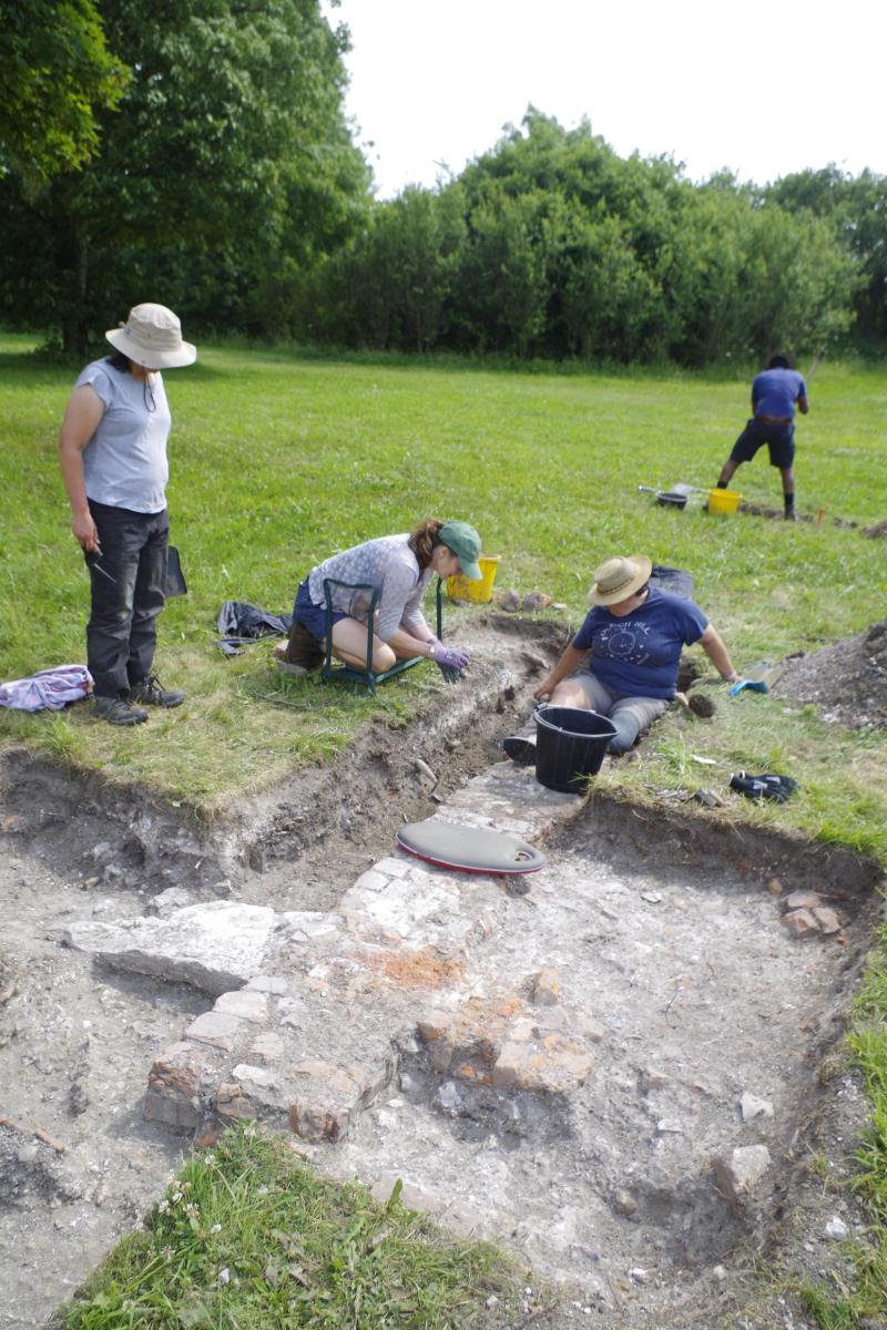 In an open field with trees, volunteers focus on the newly unearthed foundations of an old wall