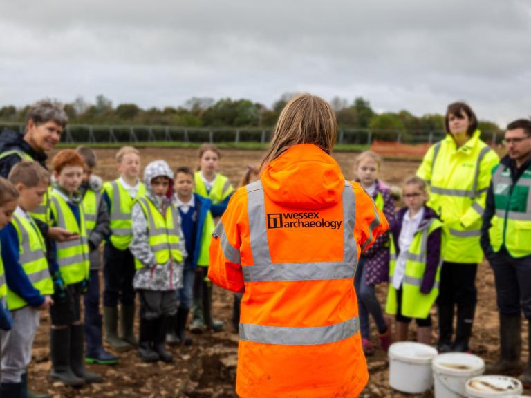 A Wessex Archaeology staff member in orange hi-vis talks to a group of school children