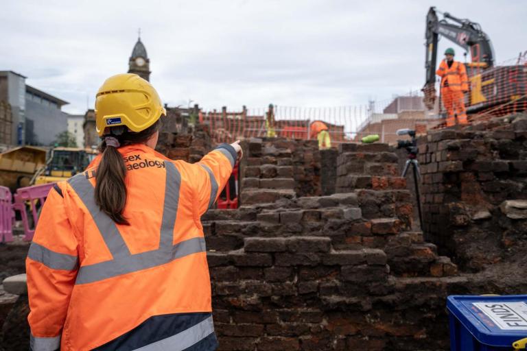 Excavations at Sheffield castle heat up with 19th c steel working discovery. With their back to the camera, an archaeologist points at brick structures in the distance