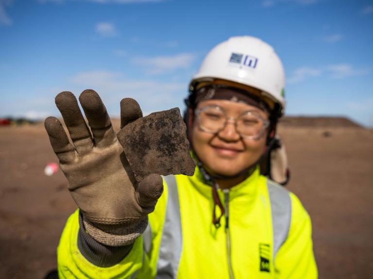 A field archaeologist holds up a black pottery sherd to the camera.
